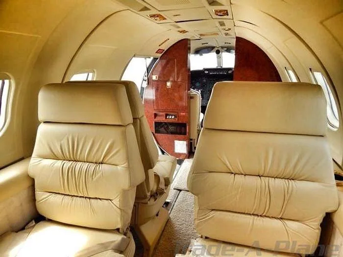 The aircraft cabin interior shows rows of light beige leather seats looking forward towards the open cockpit door.