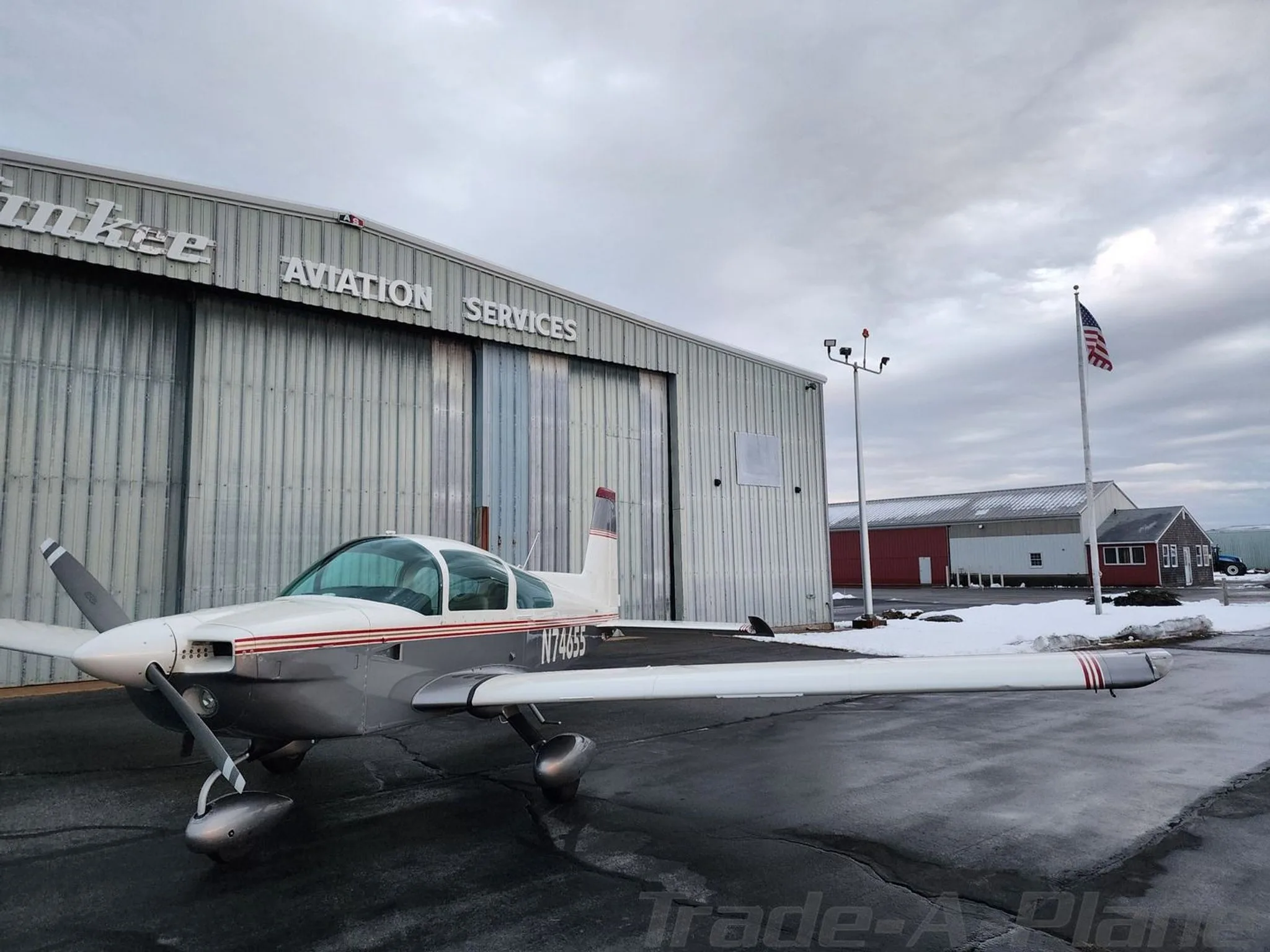A white and grey Grumman American aircraft, N74655, parked on a wet tarmac in front of a hangar with an American flag flying nearby.