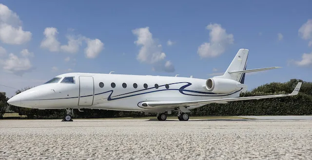 A white business jet with a blue stripe livery is parked on a tarmac under a blue sky with clouds.