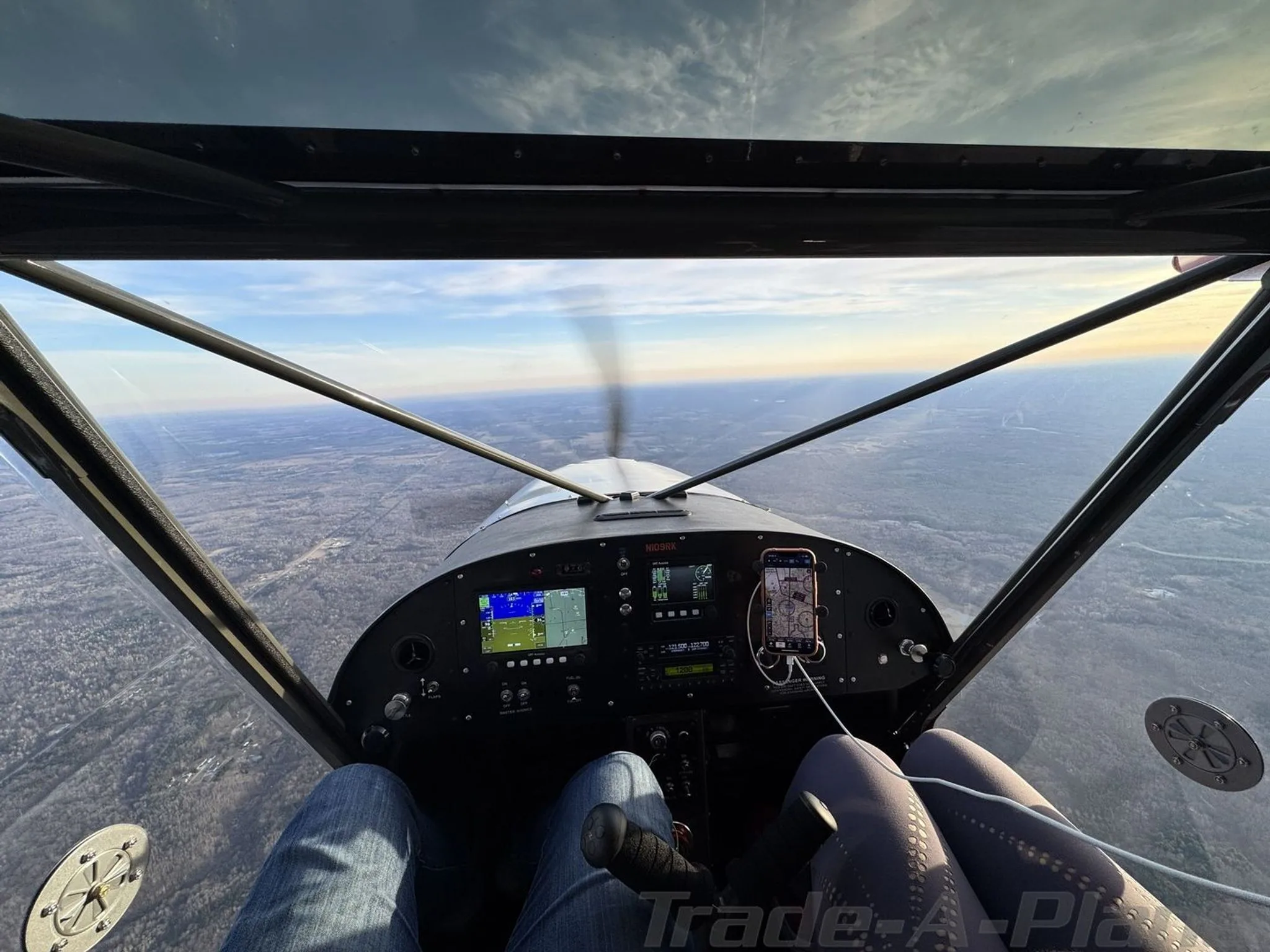 View from the cockpit of an aircraft in flight, showing the instrument panel, controls, and a pilot's legs.