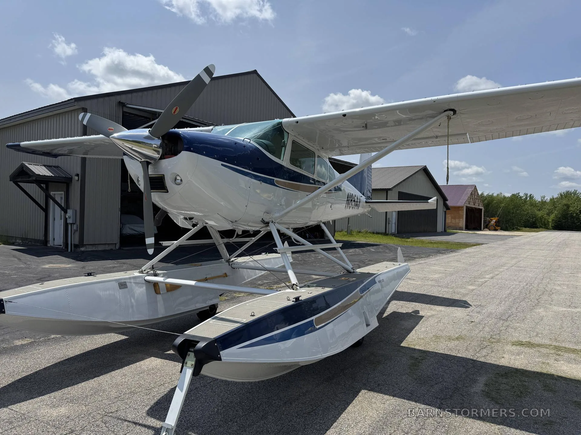 A front-quarter view of a blue and white floatplane (N85AJ) parked on an airfield with hangars under a partly cloudy sky.