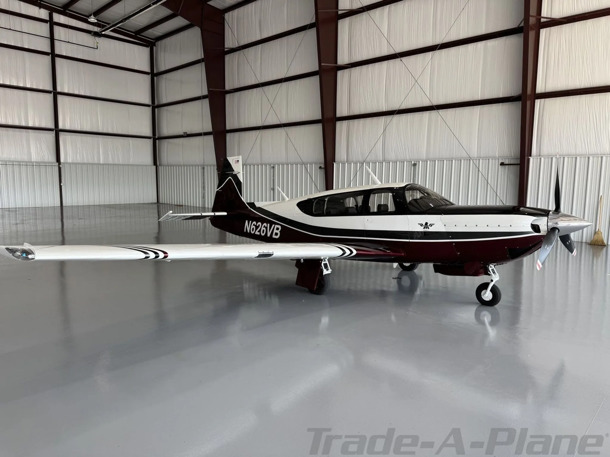 A Mooney aircraft with registration N626VB, featuring a maroon, white, and black paint scheme, is parked in a hangar, viewed from the side.