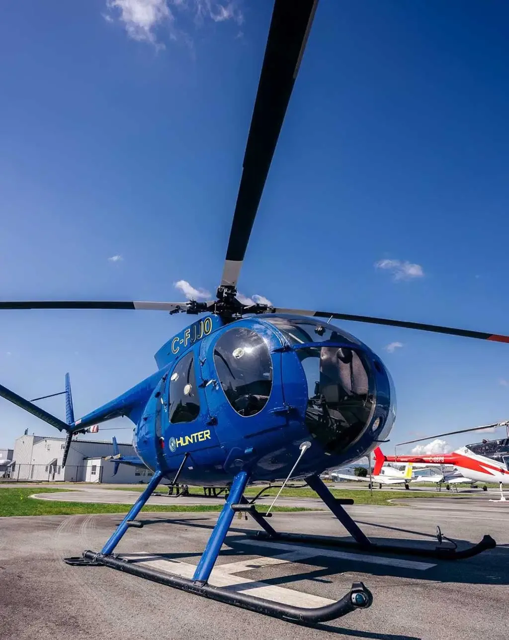 A blue helicopter with registration C-FJJ0 is parked on an airport tarmac with other aircraft in the background.