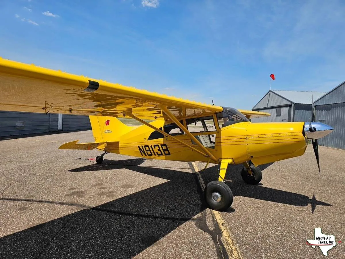 A bright yellow Maule aircraft, registration N913B, parked on an airfield tarmac with hangars in the background under a blue sky.
