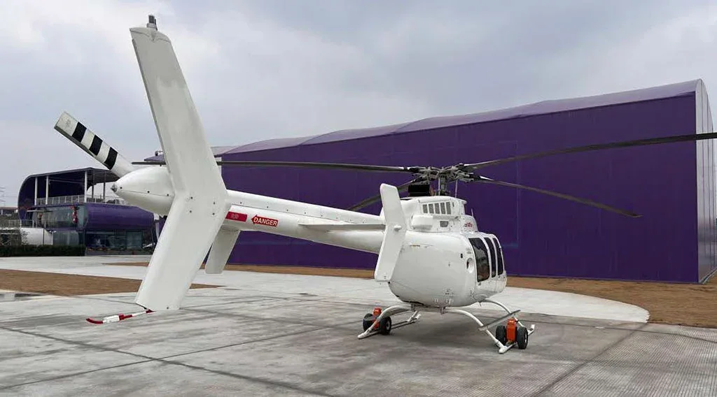 A white Bell 407GXP helicopter, viewed from the rear-right, parked on a concrete pad next to a large purple building.
