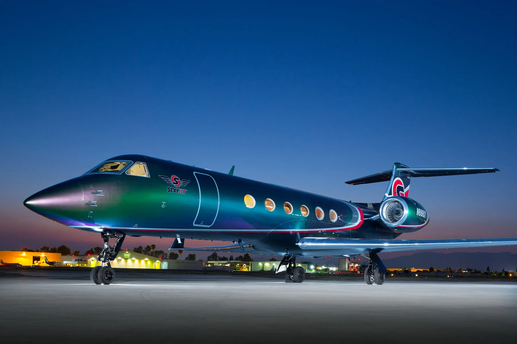 A Gulfstream business jet with an iridescent green and purple paint scheme, red accents, and 'SexyJet' logo, parked on an airfield at dusk.