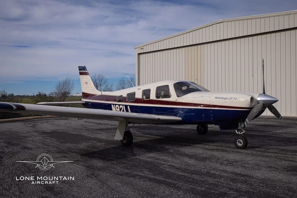A Piper Saratoga II TC aircraft (N92LL) with blue and white paint scheme parked on an asphalt apron in front of a hangar.