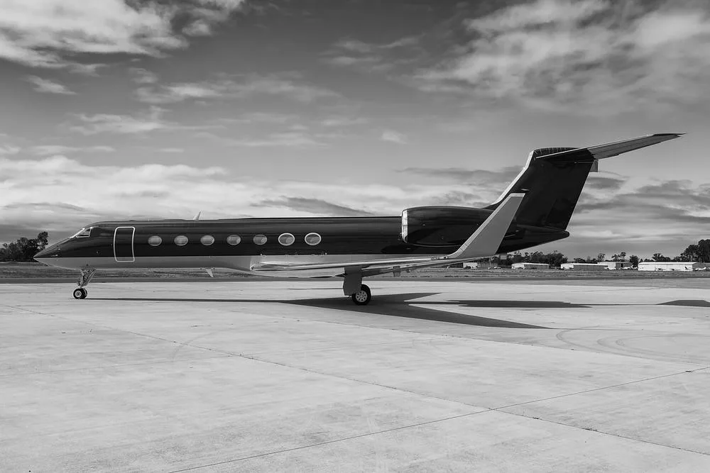 A black and white side view of a sleek business jet, likely a Gulfstream G650, parked on an airfield under a cloudy sky.