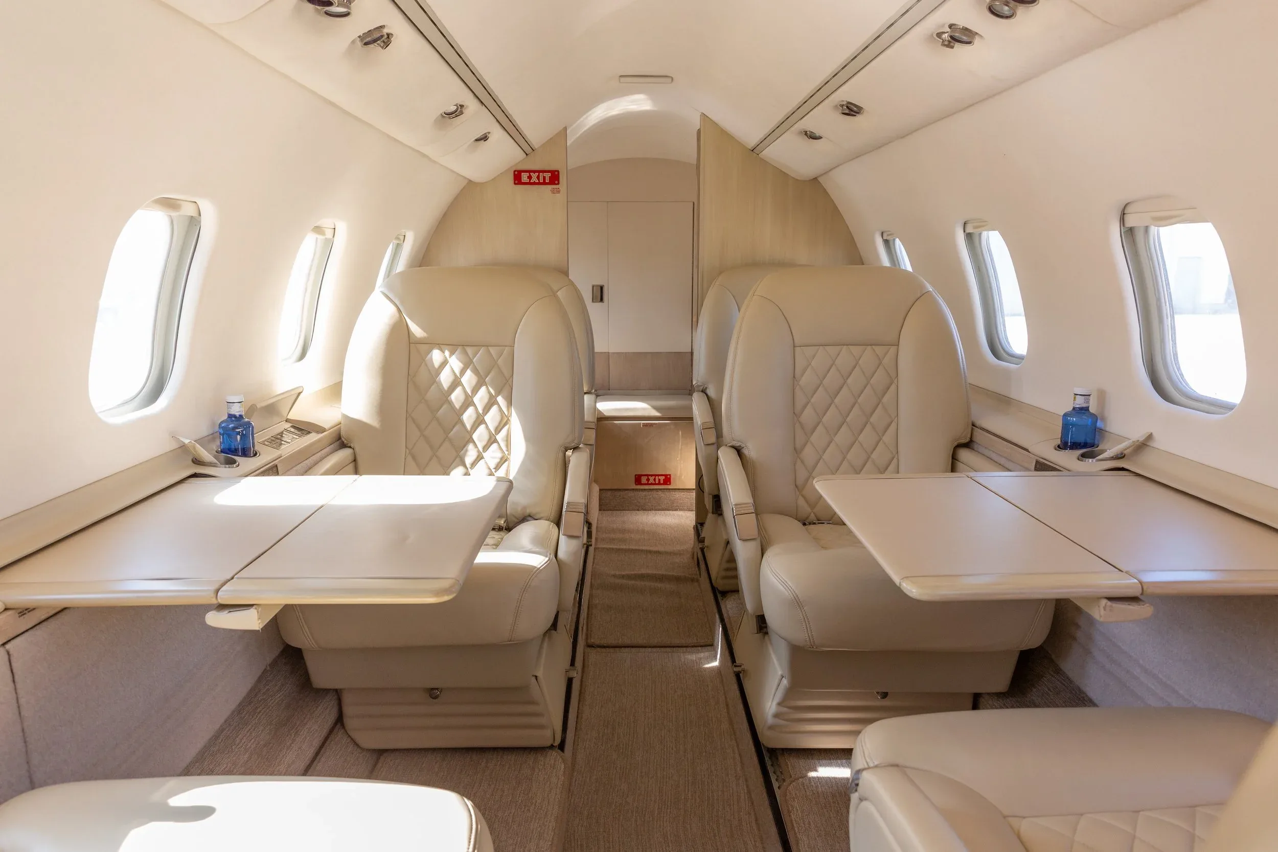 The interior cabin of a private jet with light-colored quilted leather seats and fold-out tables.