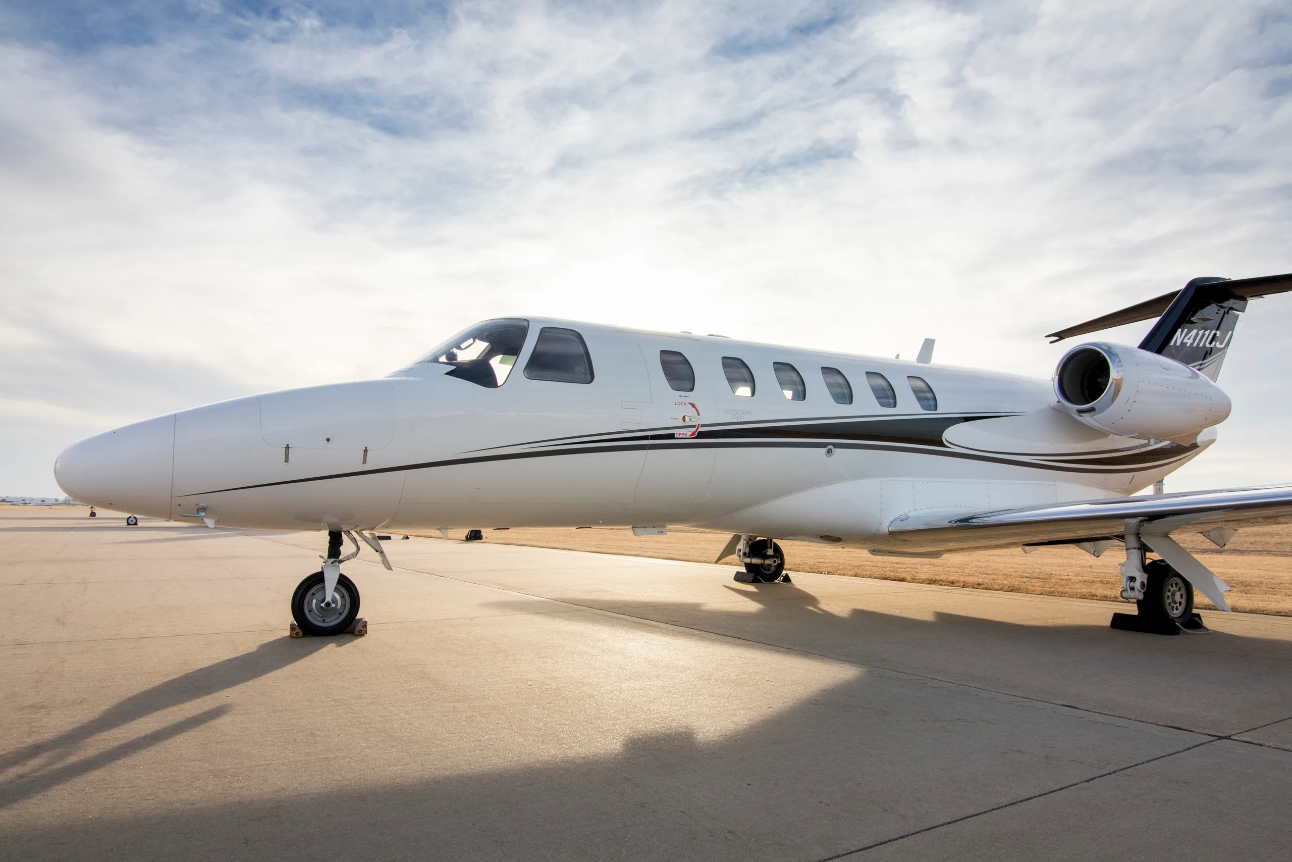 A white Cessna Citation CJ3 private jet with black and gold stripes on the tail, registration N411CJ, parked on an airfield with the sun low in the sky.