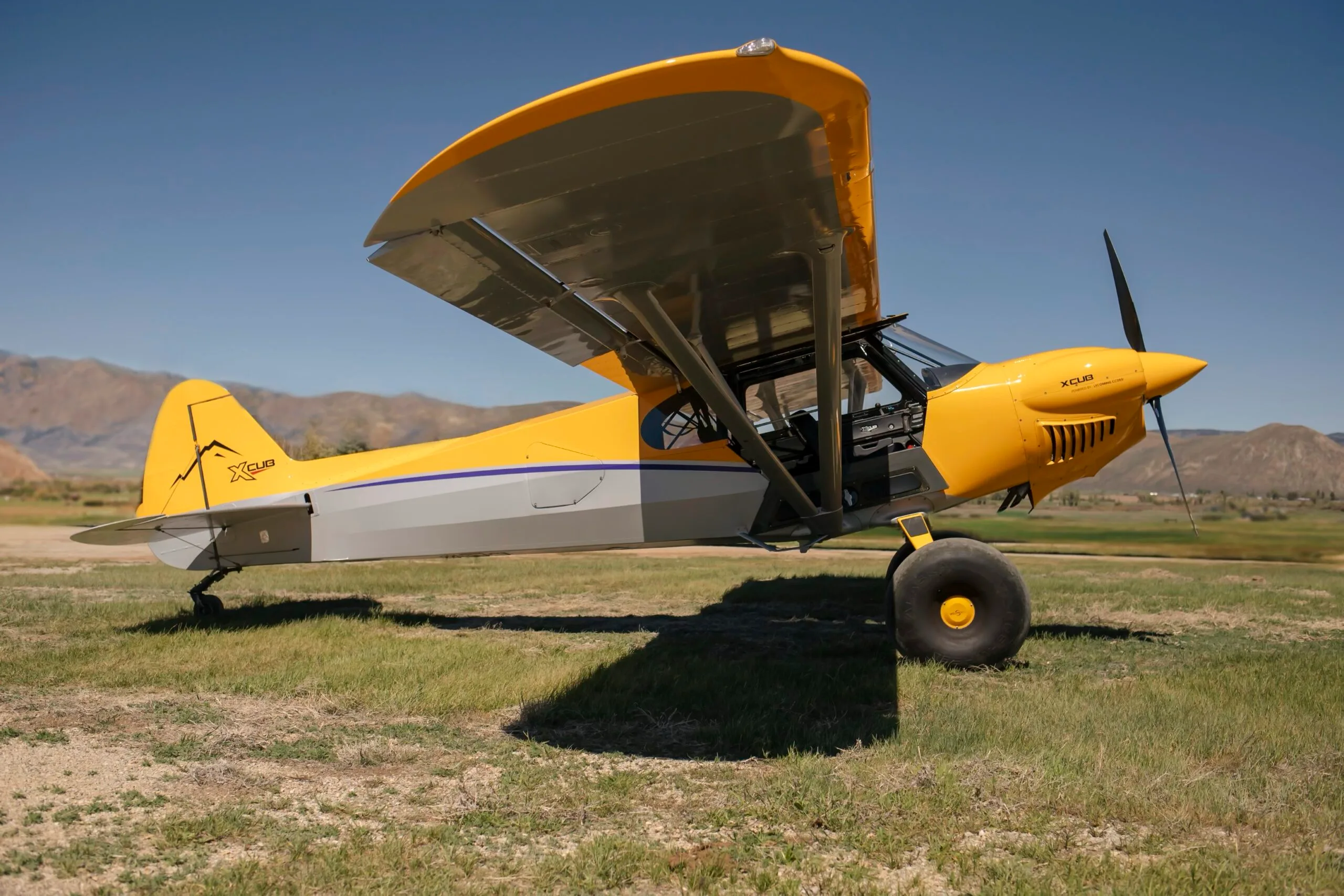 A side view of a yellow and silver CubCrafters XCub aircraft with large bush tires on a grassy field.