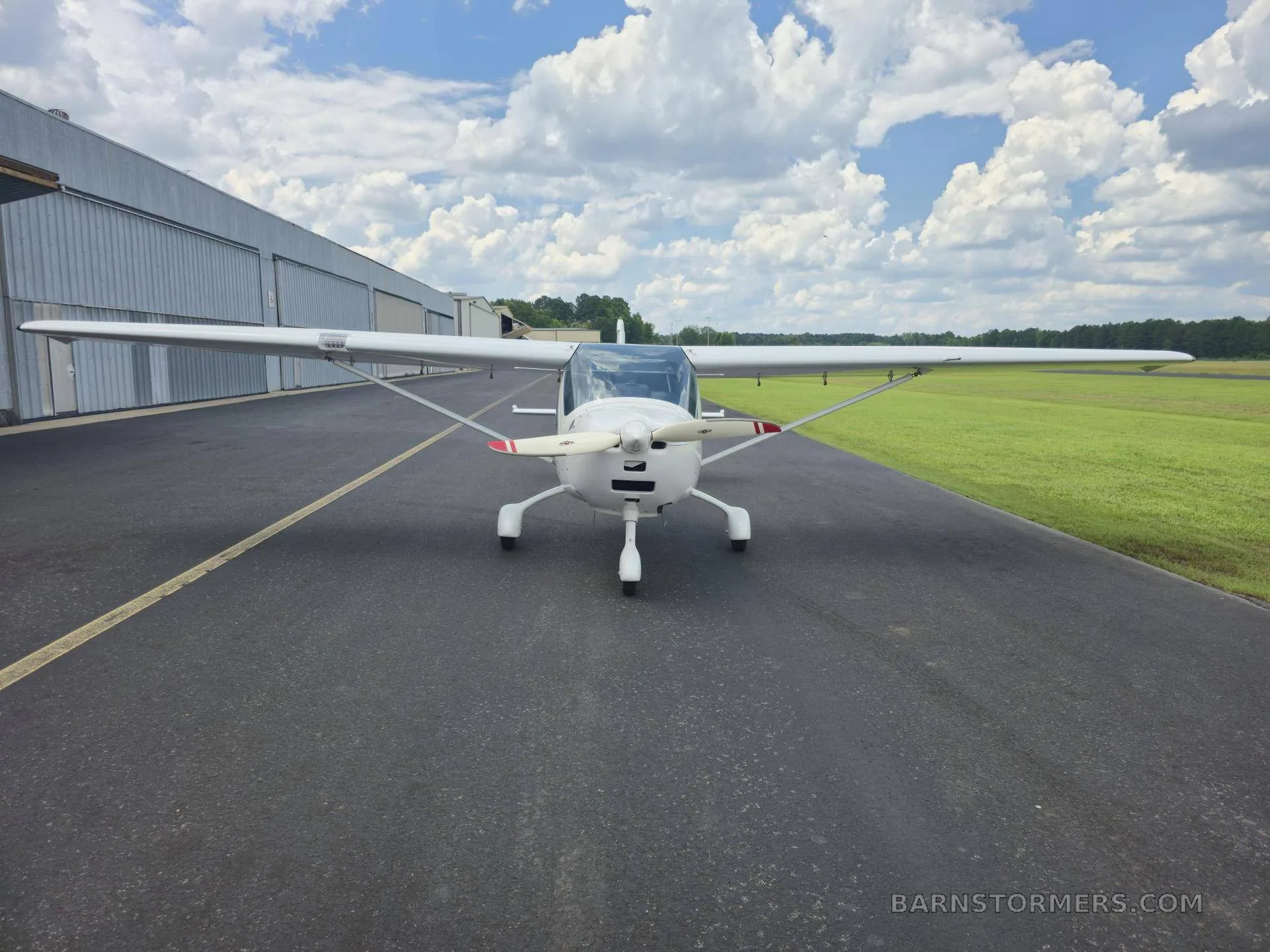 A white Remus GX Light Sport aircraft is viewed head-on, parked on an asphalt runway in front of a hangar.