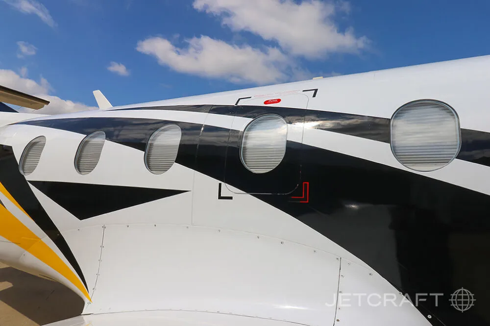 A close-up view of the white, black, and yellow livery on the fuselage of a Pilatus PC-12 aircraft, showing passenger windows.