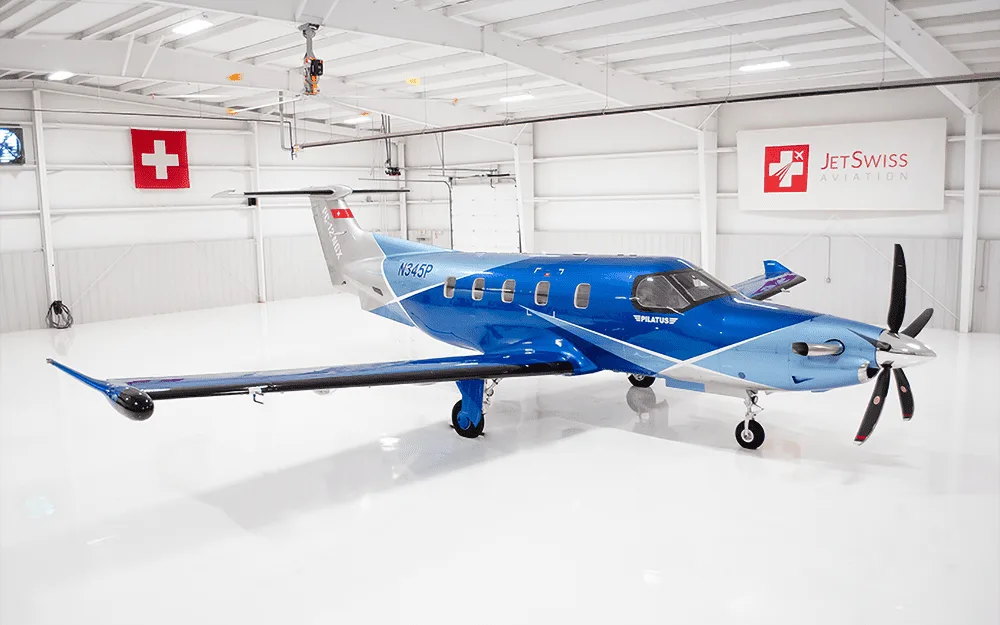 A Pilatus PC-12 NGX aircraft with a blue and silver paint scheme, registration N345P, parked inside a white hangar.
