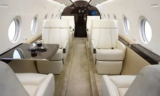 A private jet cabin interior featuring cream leather seats, a central aisle, and a table with drinks.