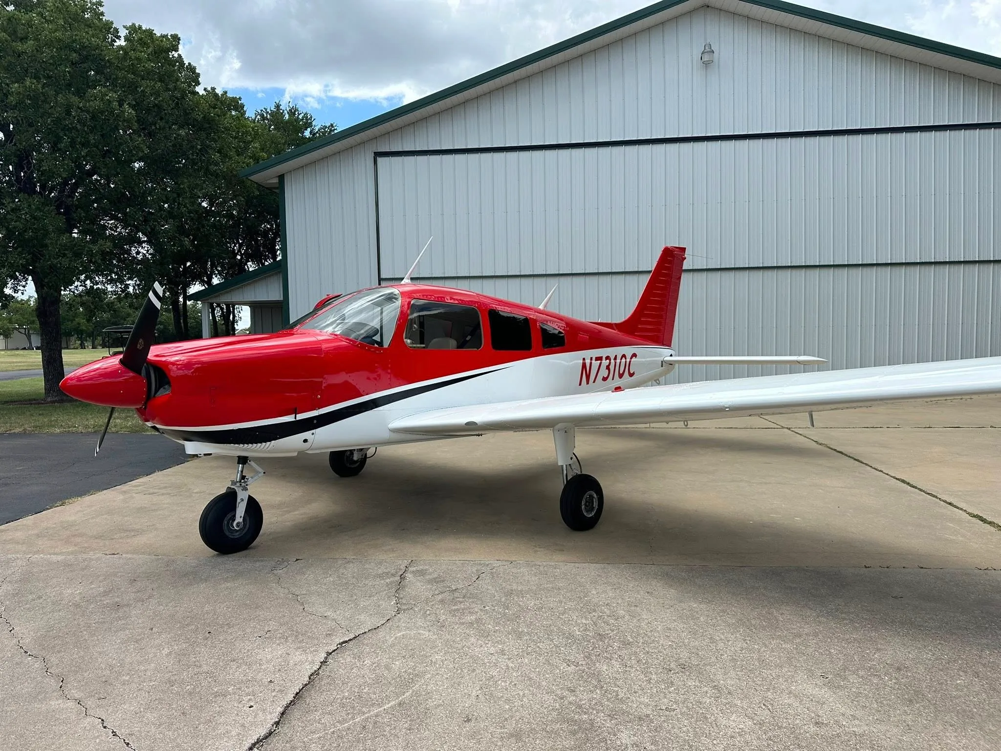 A red and white single-engine propeller aircraft, possibly a Piper Cherokee, parked on a concrete apron in front of a white hangar.
