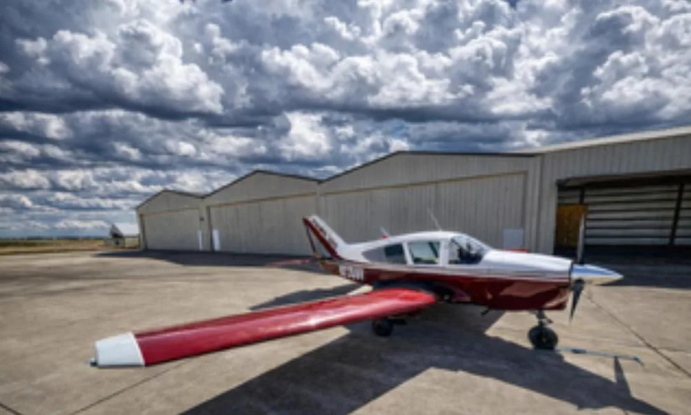 A red and white Bellanca Viking 300 (N7356V) parked on an airfield in front of hangars under a dramatic cloudy sky.