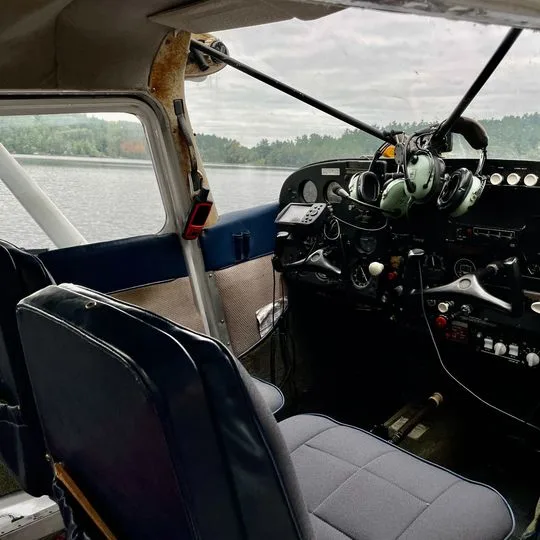 The interior of a floatplane shows the cockpit with instruments and controls, and two grey seats, looking out over water.
