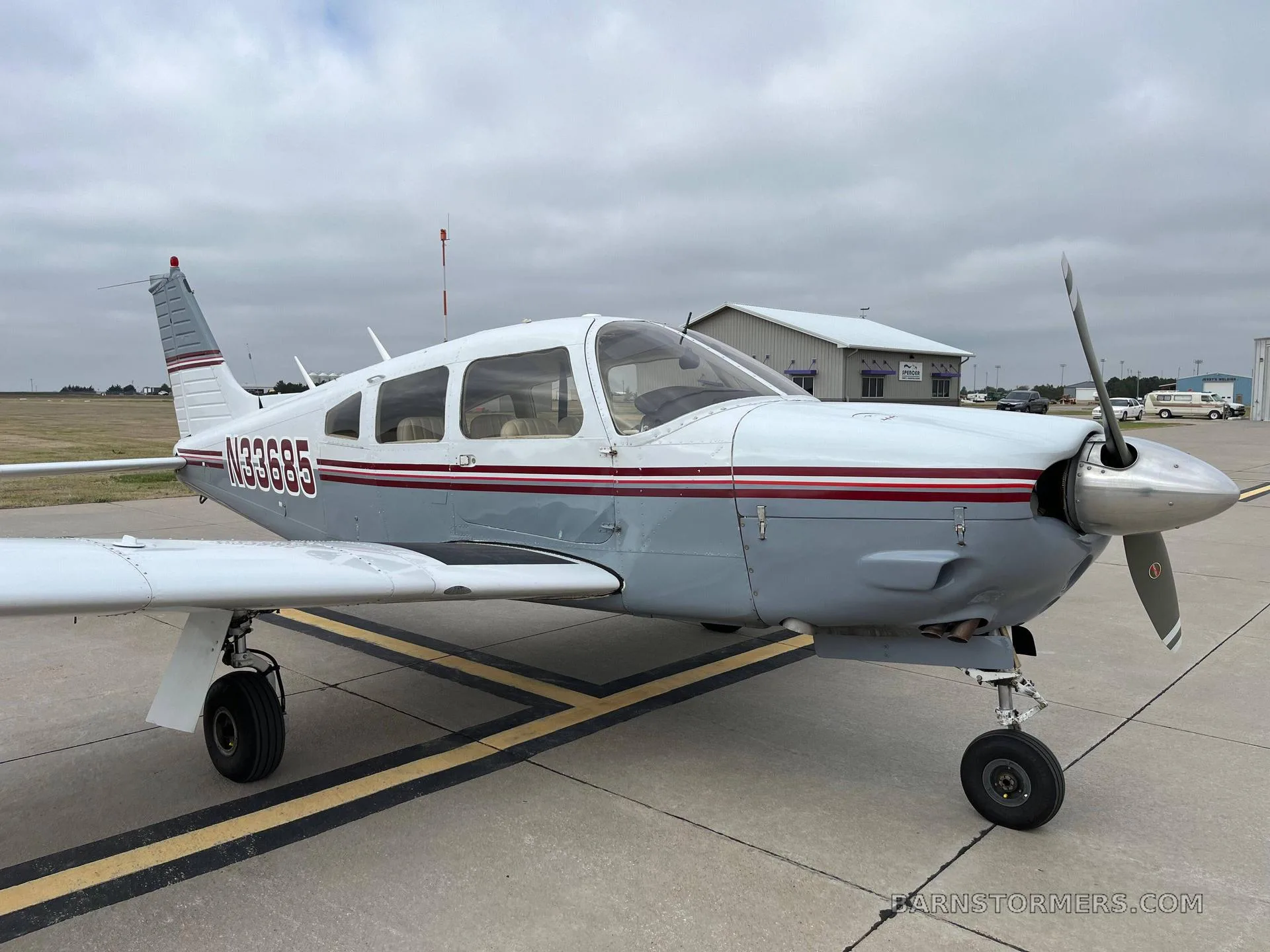 A light gray and white single-engine aircraft with a red stripe, registration N33665, parked on an asphalt ramp under a cloudy sky.