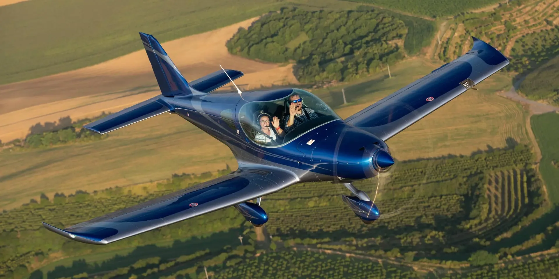 A sleek, dark blue single-engine aircraft with two people waving from the cockpit flies over green and brown agricultural fields.