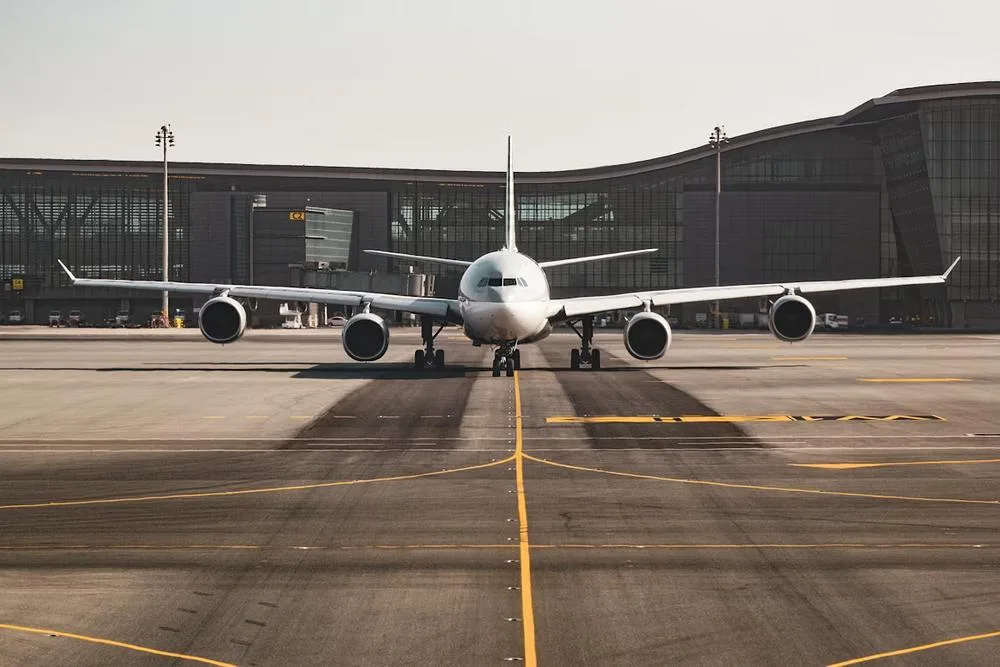 A white wide-body aircraft, likely an Airbus A330, is seen head-on on an airport tarmac with a modern terminal building in the background.