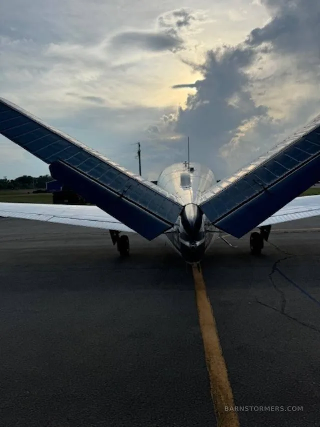 A rear view of a white and blue V-tail aircraft with its wings folded up, on an asphalt runway under a cloudy sky.