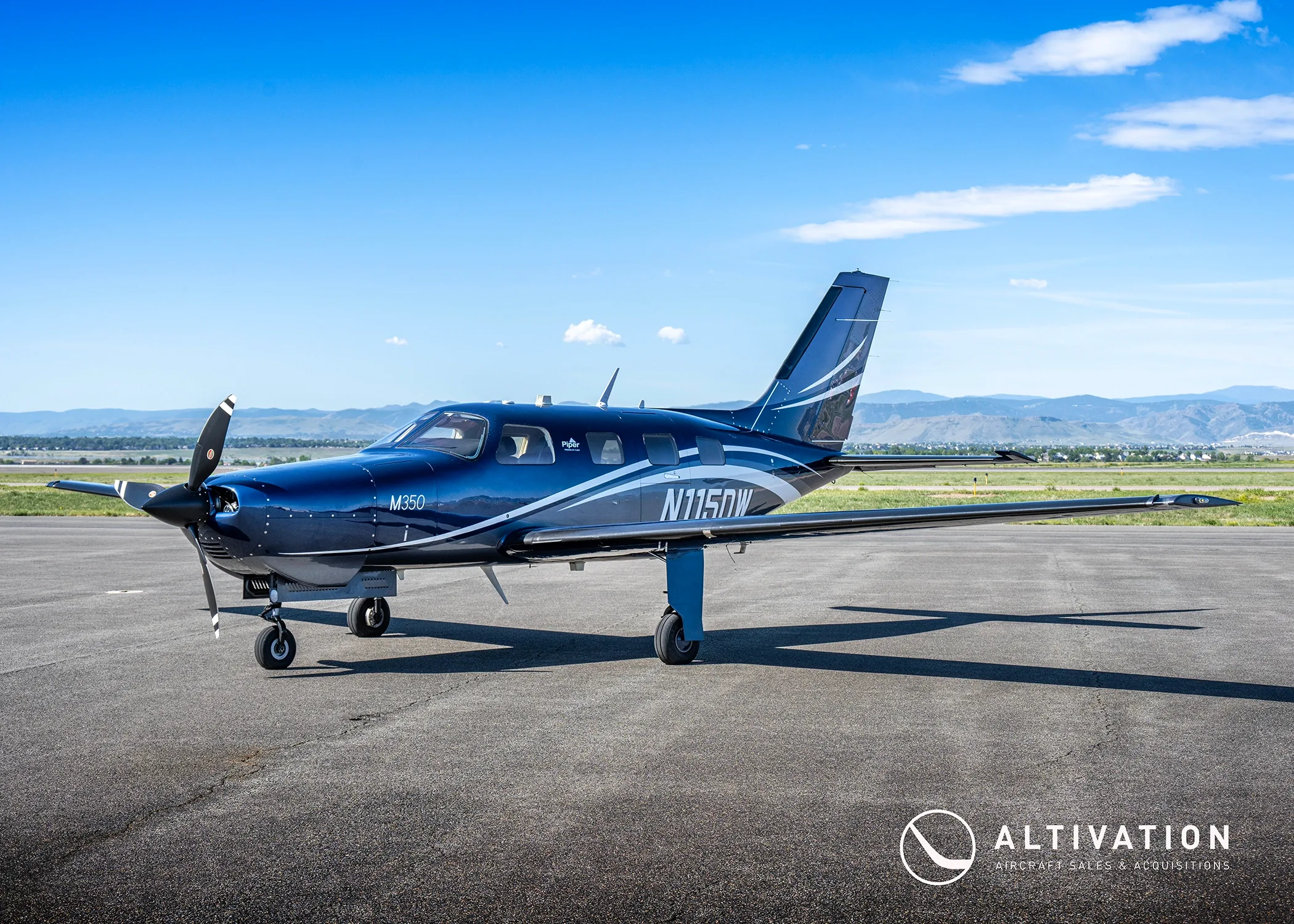 A dark blue Piper M350, tail number N115DW, parked on a tarmac with a clear blue sky and distant mountains.