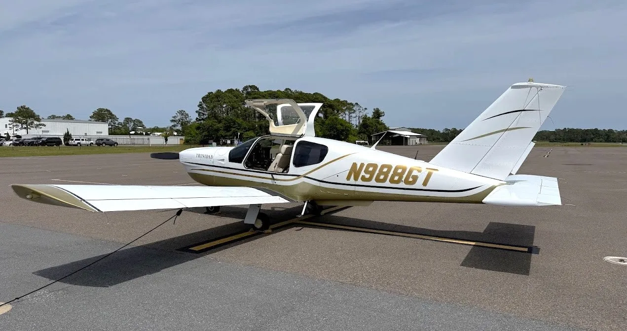 A white and gold Socata TB-20 Trinidad aircraft with its doors open is parked on an airfield.