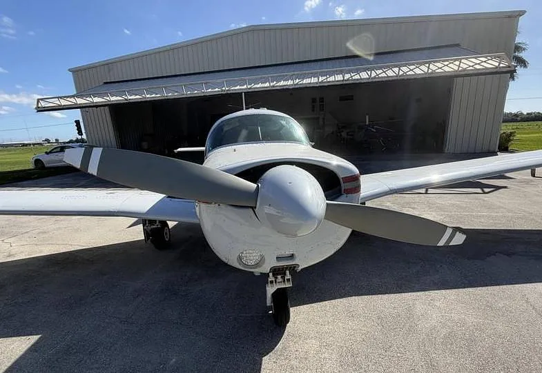 Front view of a white single-engine aircraft with a two-blade propeller, parked in front of a hangar.