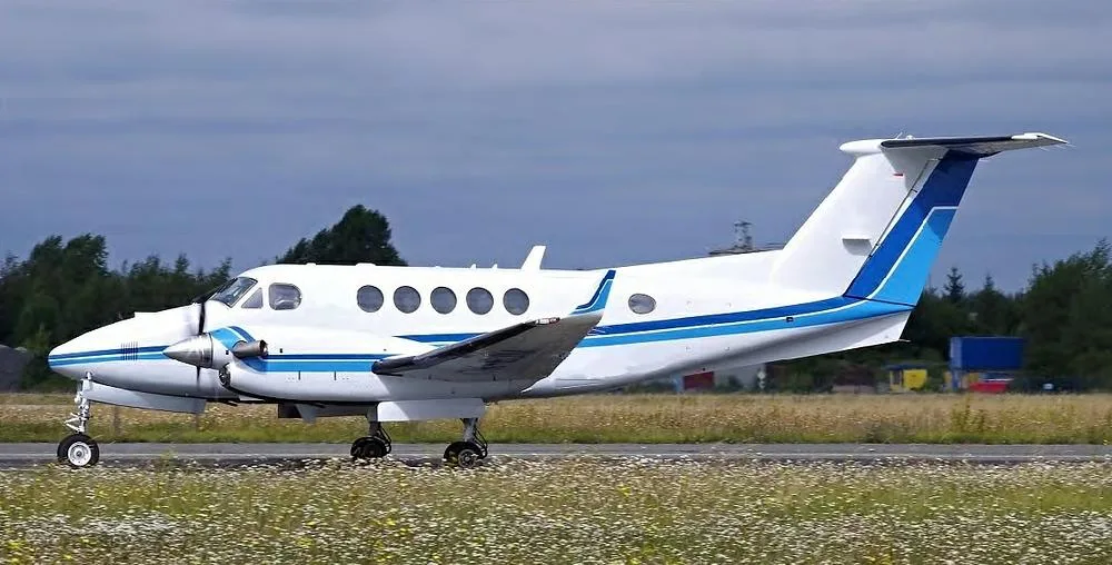 A white twin-turboprop aircraft with blue stripes on the fuselage and tail, on a runway with grass and trees in the background.