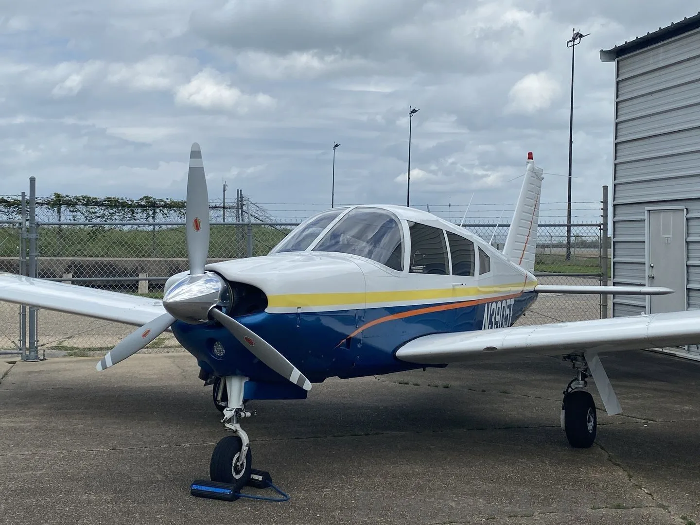 A Piper Cherokee-style aircraft, N3905T, with a white, blue, and yellow paint scheme, parked on a concrete apron next to a hangar under a cloudy sky.