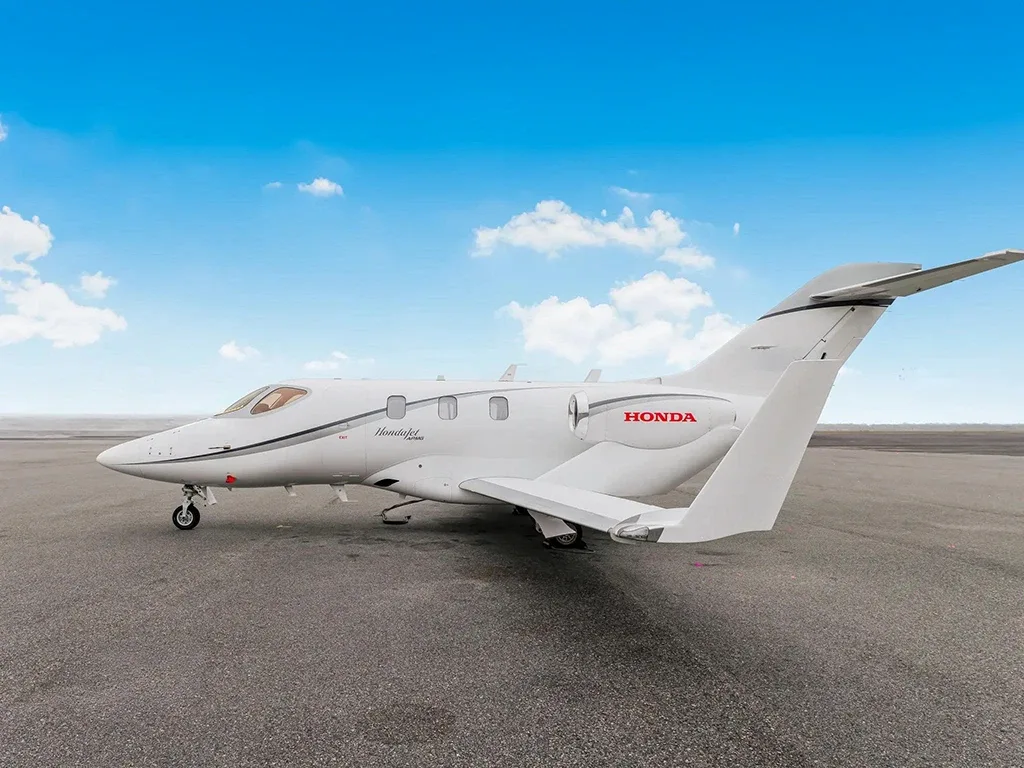 A white HondaJet HA-420 business jet with red 'HONDA' branding is parked on an asphalt tarmac under a blue sky.