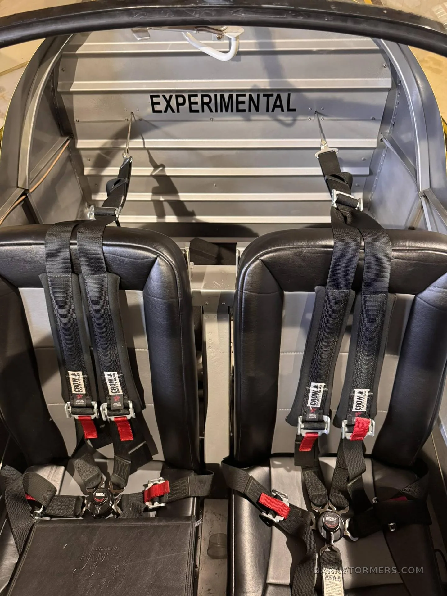 The interior of an experimental aircraft shows two black seats with multi-point harnesses and "EXPERIMENTAL" stenciled on the bulkhead.