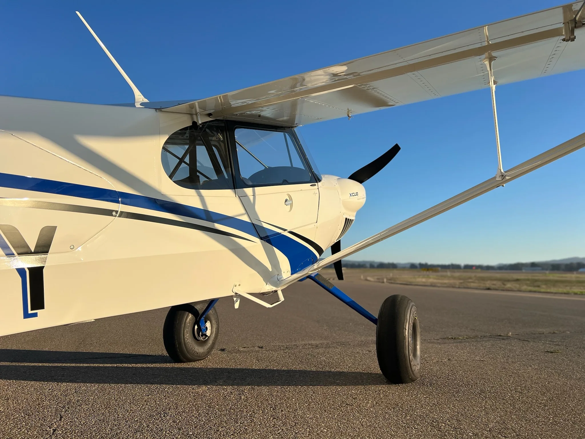 A close-up view of the front-right side of a white X-Cub aircraft with blue accents, showing the propeller and landing gear.