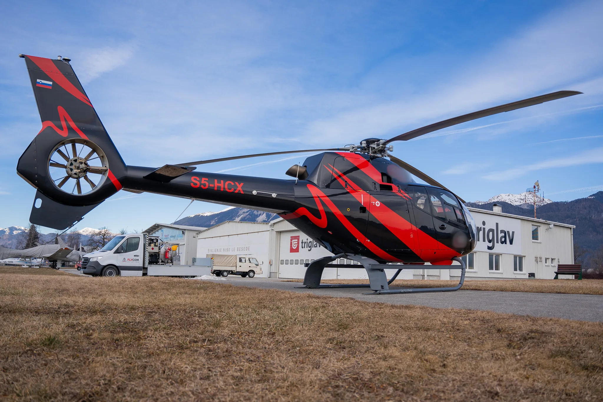 The S5-HCX helicopter is parked outside a building with the Triglav logo.