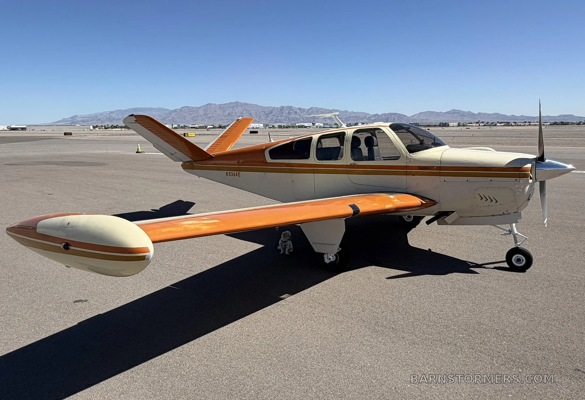 A cream and orange single-engine aircraft with tail number N5364E parked on an airfield with mountains in the background.