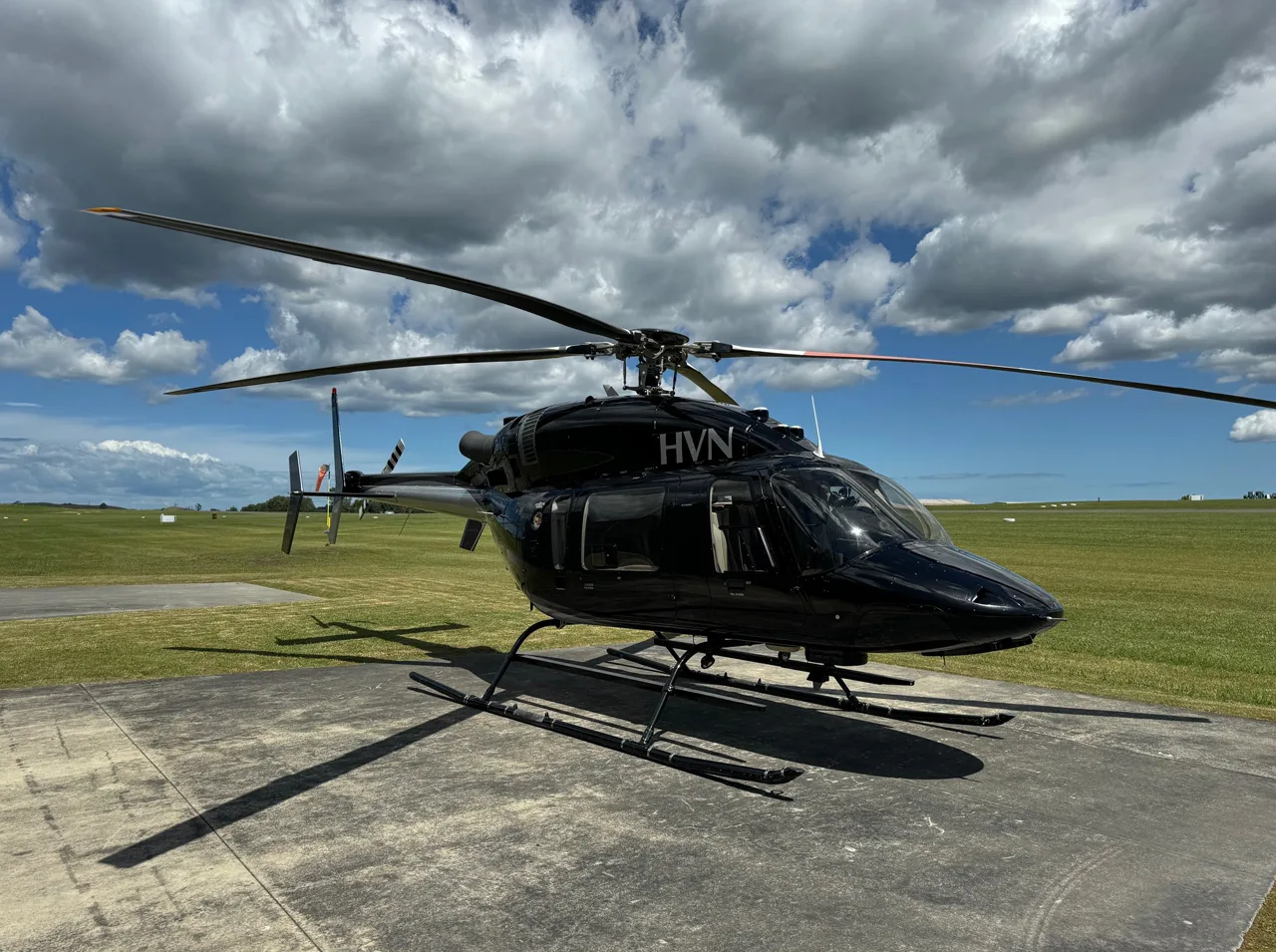 A black helicopter with 'HVN' registration is parked on a concrete pad at an airfield under a partly cloudy sky.