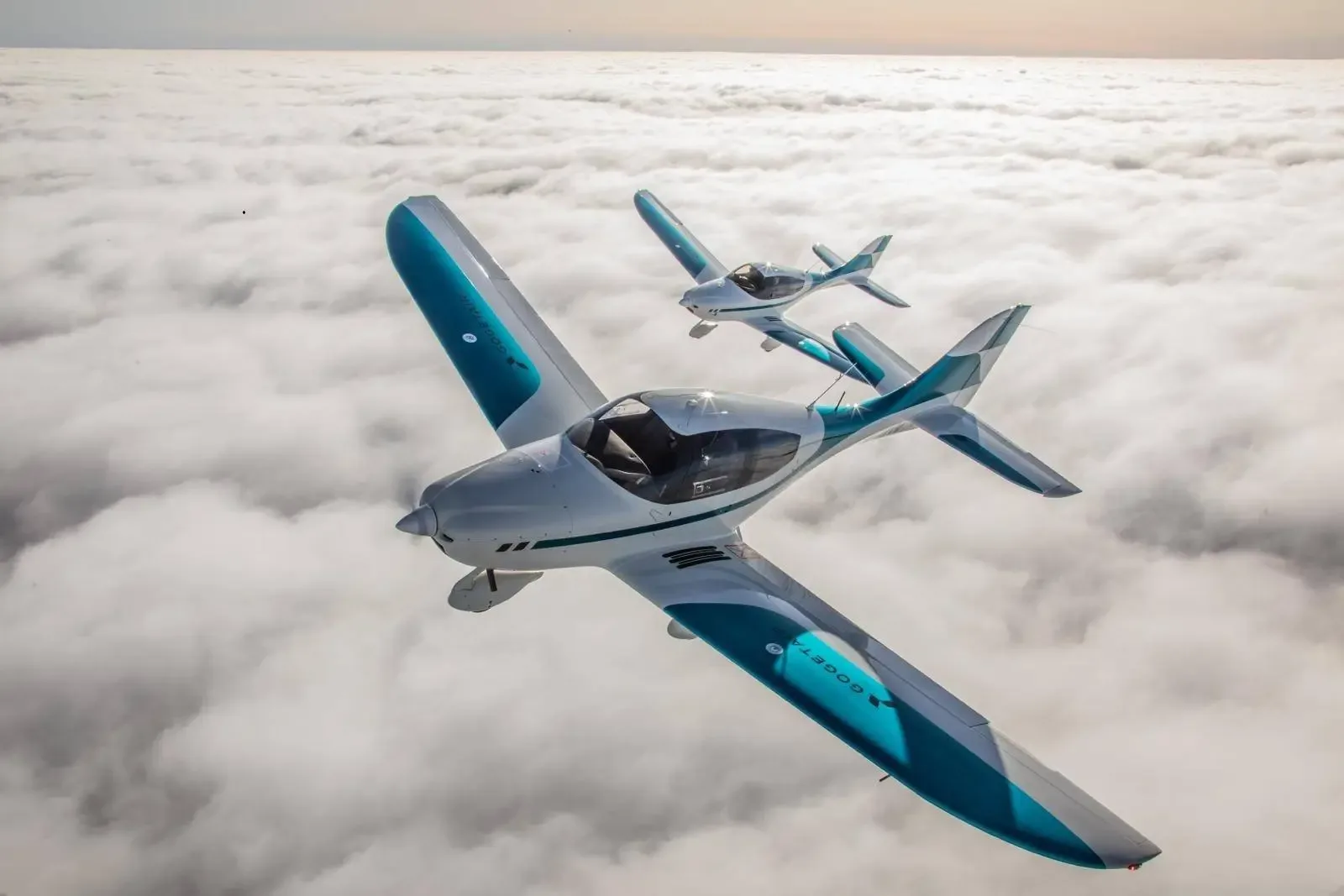 Two white and teal light aircraft fly in formation above a thick layer of clouds.