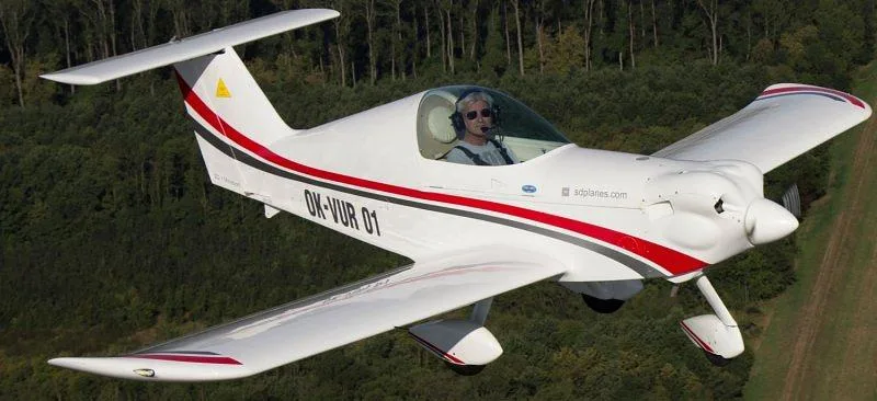 A white light sport aircraft with red and gray stripes, registration OK-VUR 01, flying over a green forest.