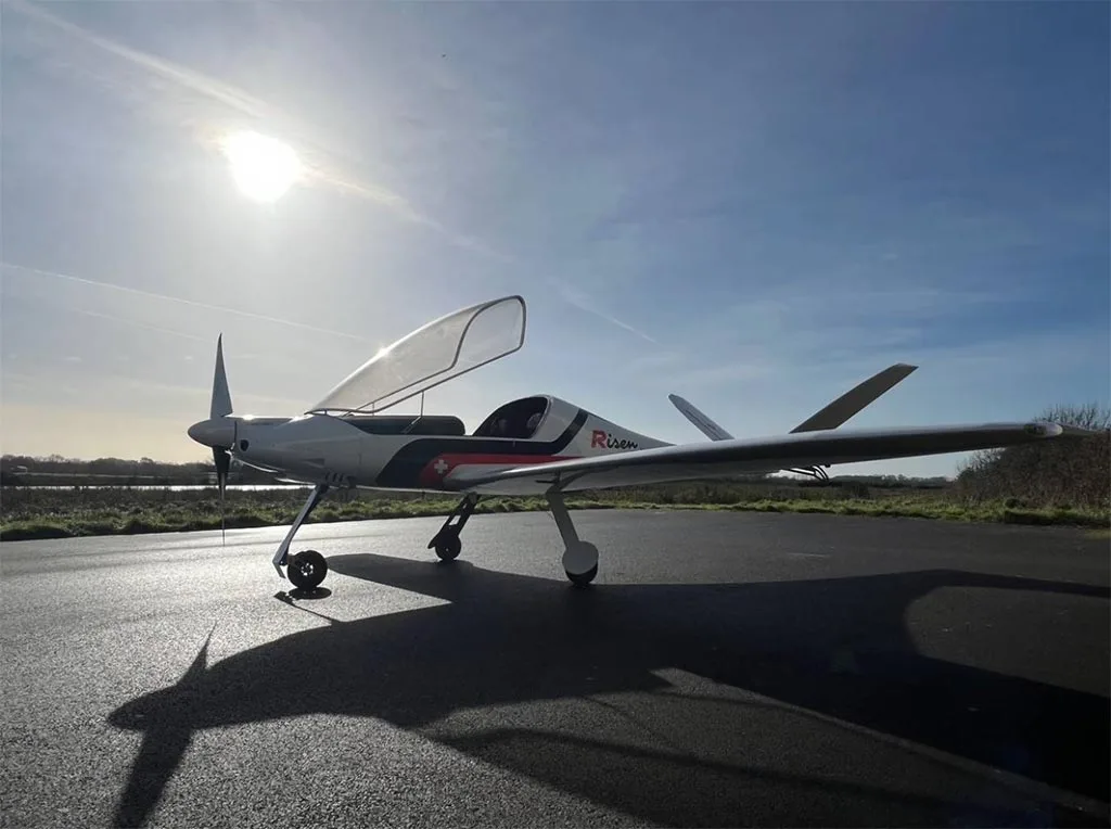 A white Risen aircraft with an open canopy is parked on an airfield with the sun low in the sky.