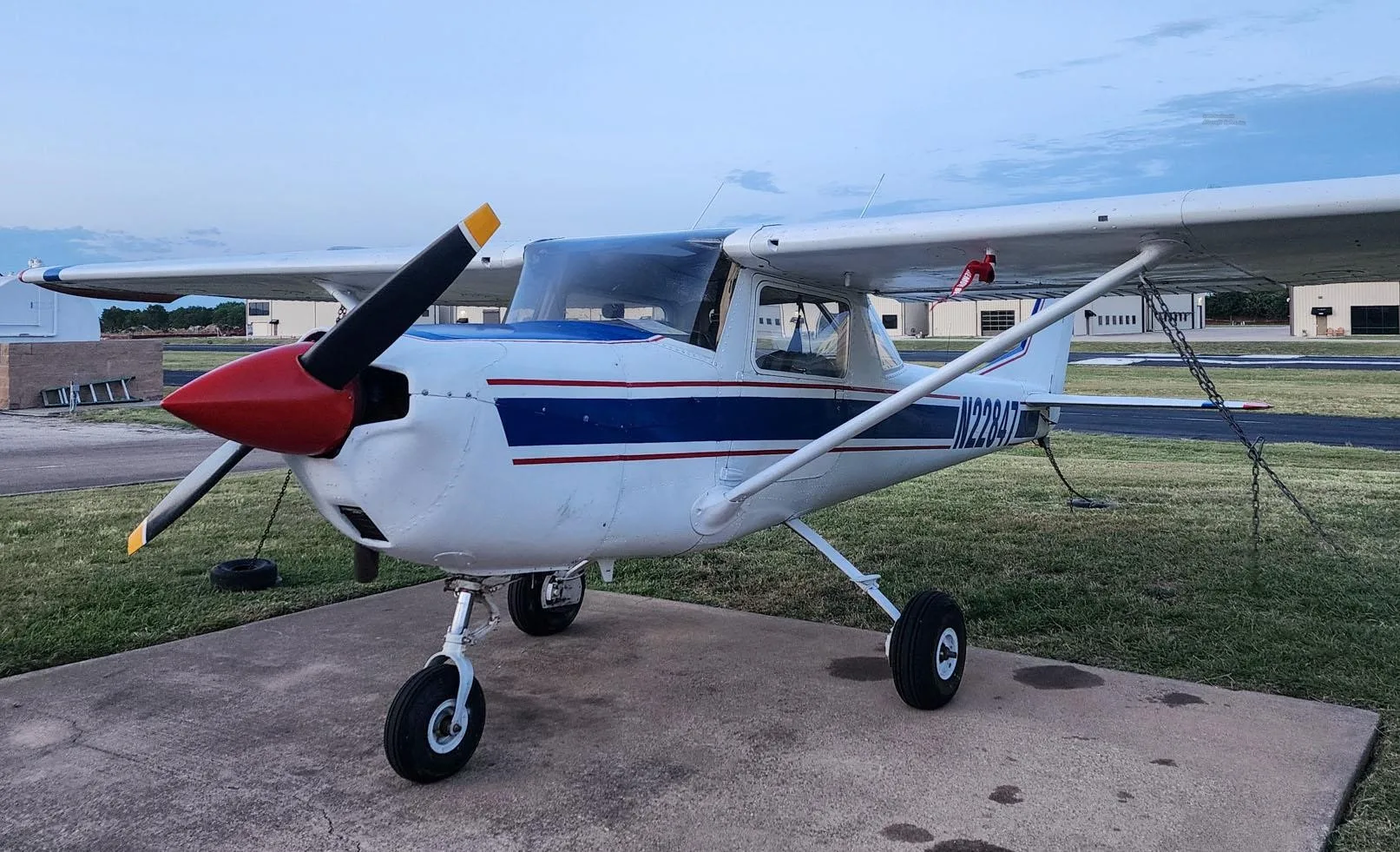 Three-quarter front view of a white Cessna 150 with blue stripes and a red propeller spinner, parked on a concrete pad.