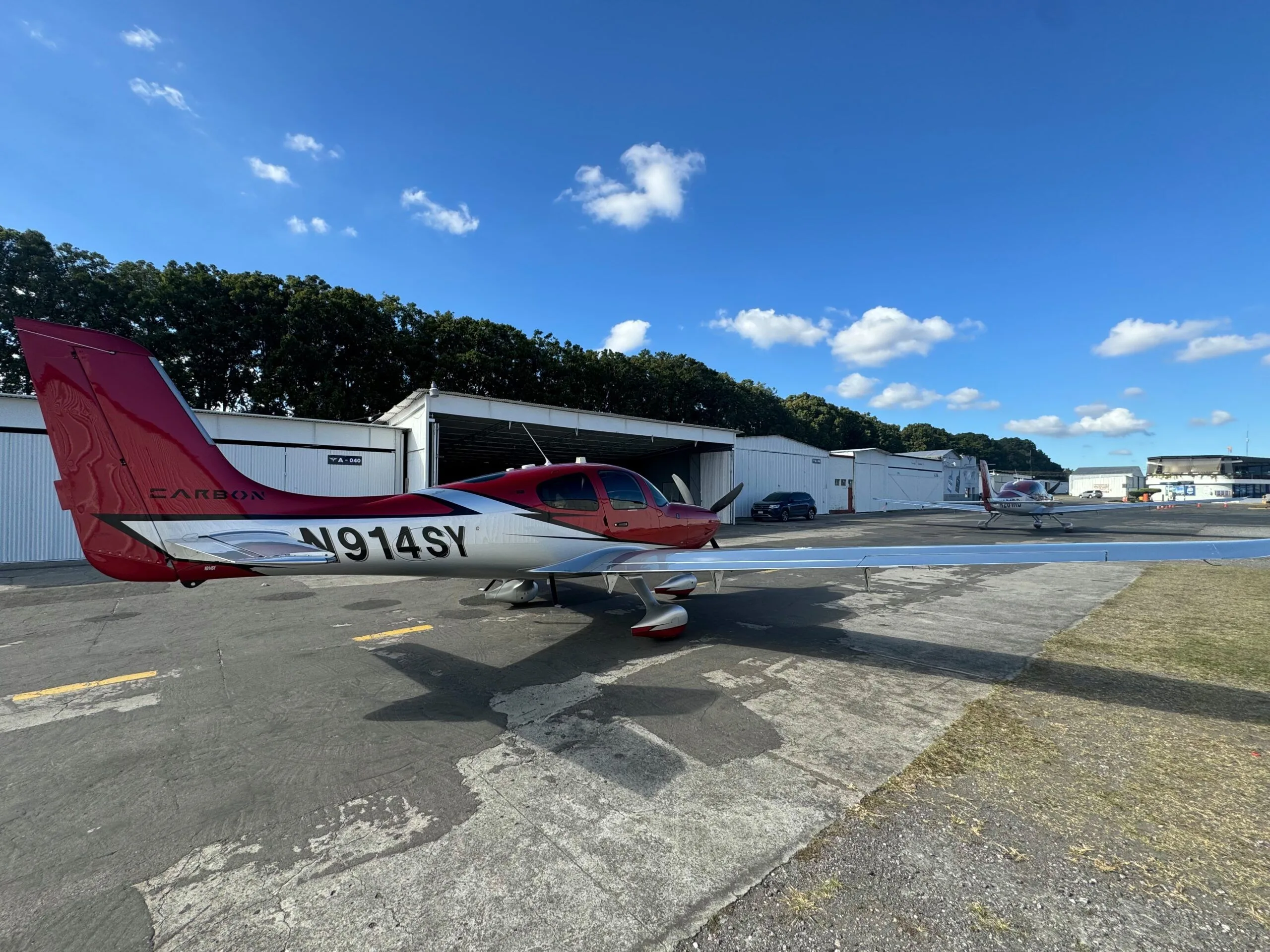 The red and silver Cirrus SR22 aircraft N914SY parked on an airfield with hangars and trees in the background.