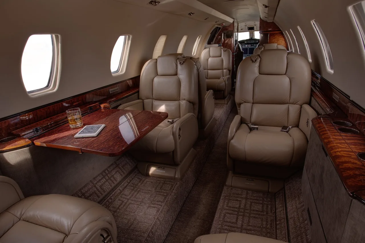 Aircraft interior with leather seats, wood accents, and a table with a drink.