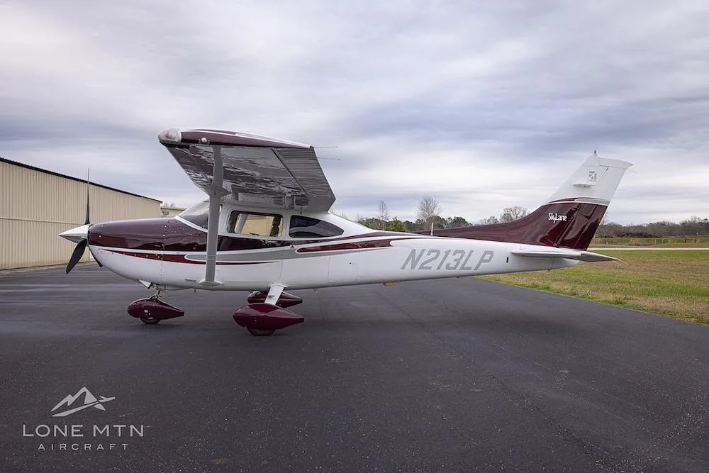 A Cessna 182 Skylane with a maroon and white paint scheme, parked on an asphalt ramp under a cloudy sky, viewed from the left side.