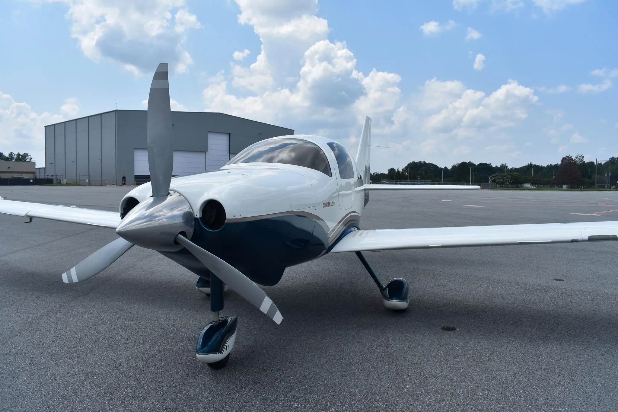 A front view of the white and blue Columbia 400 aircraft, registration N371BG, parked on an airfield with a hangar in the background.