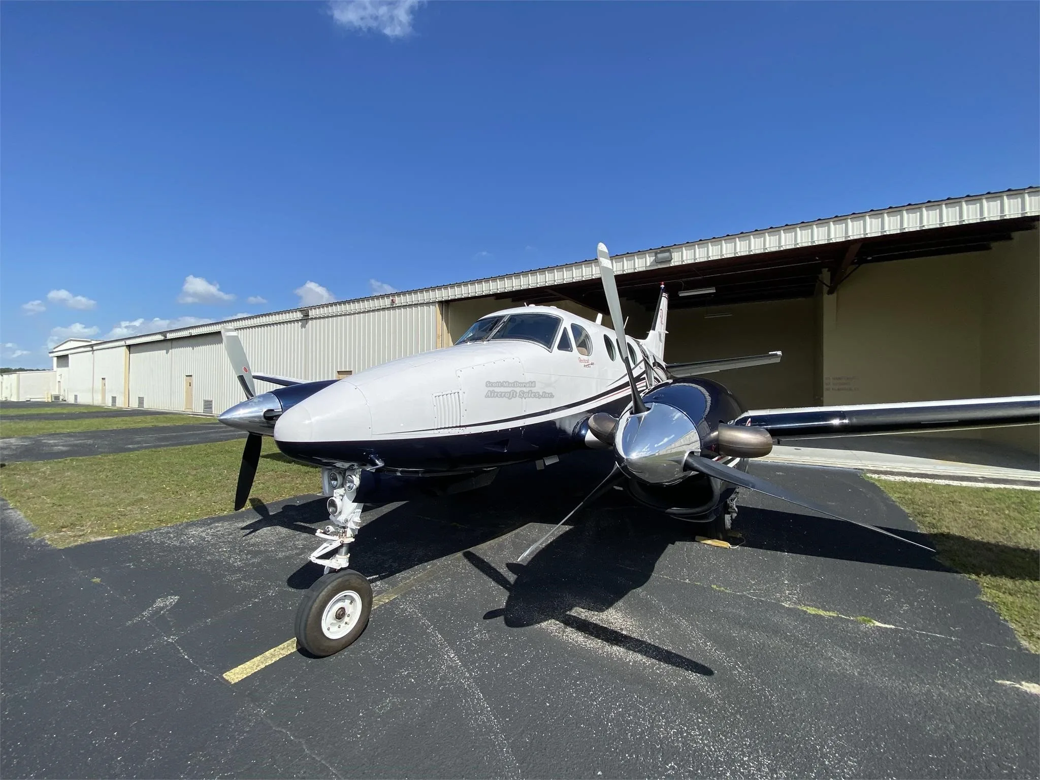 A head-on view of a white and dark blue Beechcraft King Air with its propellers, parked on an asphalt apron.