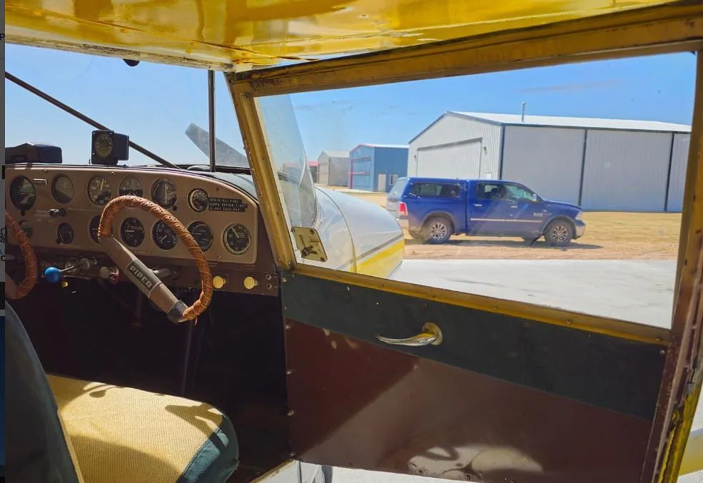 View from inside the cockpit of a Piper PA-12 Super Cruiser showing the instrument panel and a blue truck outside.