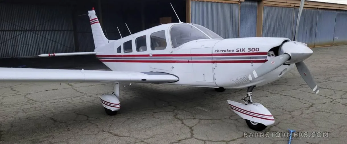 A white Piper Cherokee Six 300 aircraft with red and grey stripes, seen from the front-right, parked on an asphalt apron in front of a hangar.