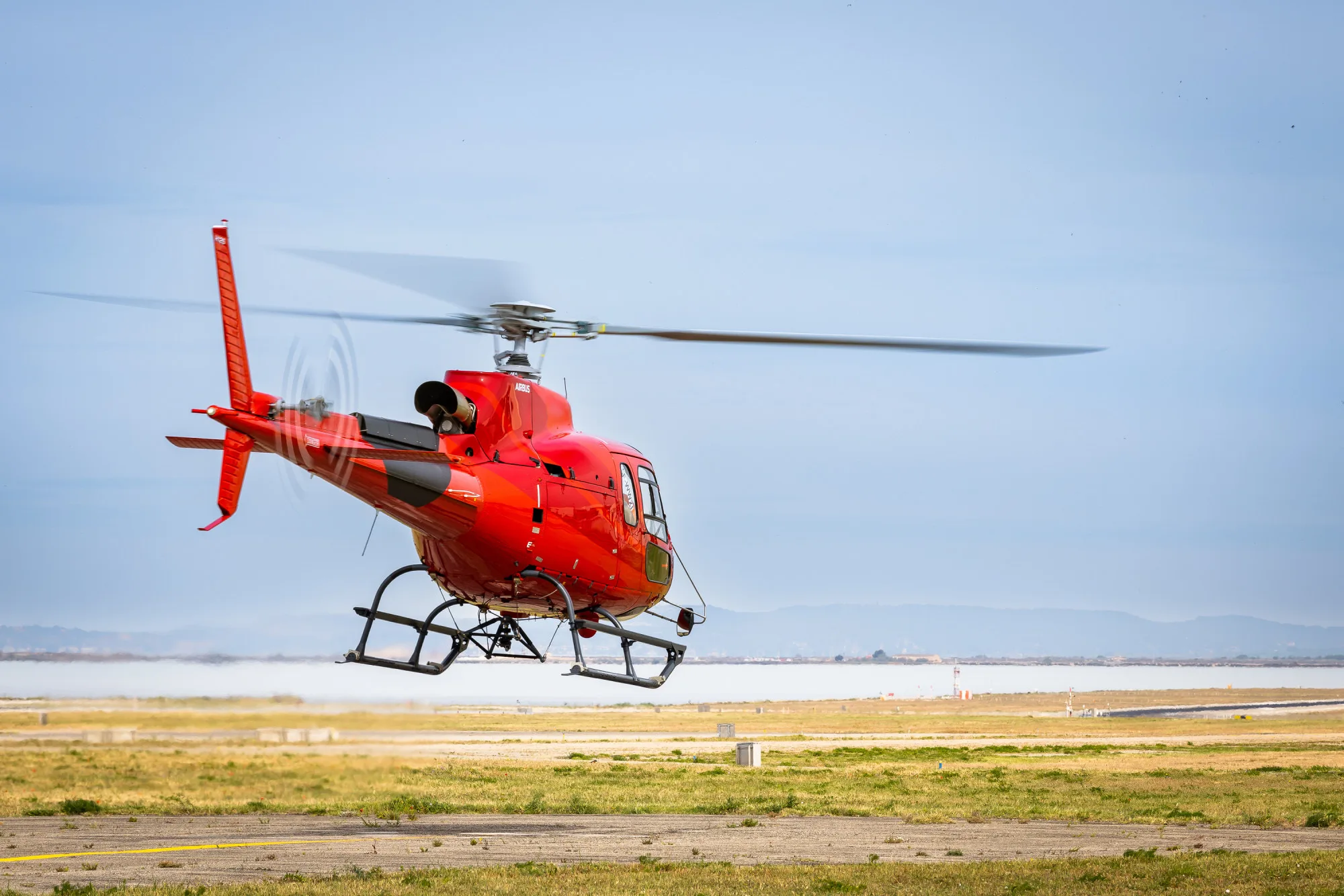 A red Airbus H125 helicopter is captured mid-takeoff from a grassy airfield, with a body of water and distant hills in the background.
