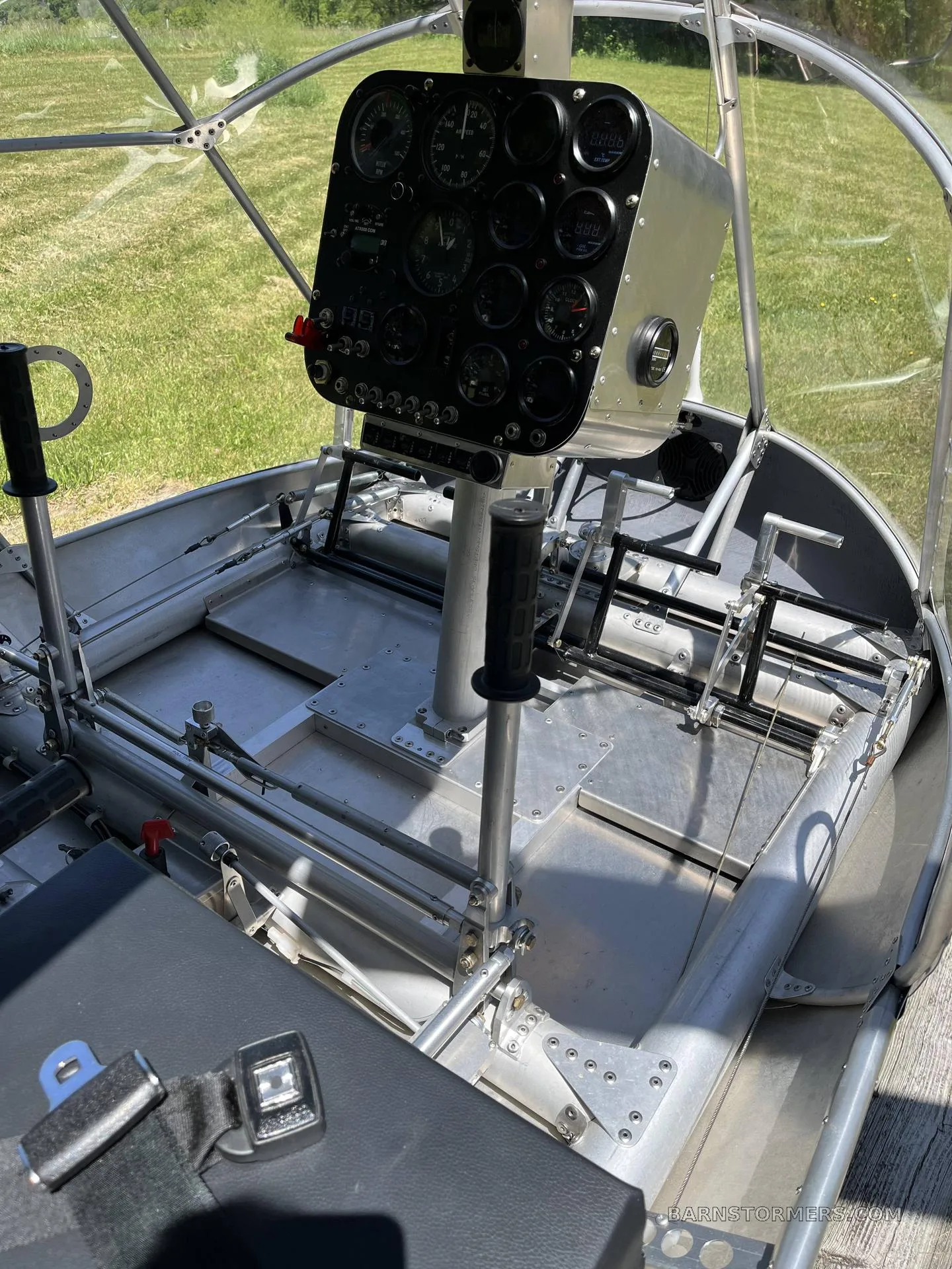 The interior of a single-seat ultralight aircraft, showing the black instrument panel, control sticks, and a black seat with a seatbelt.